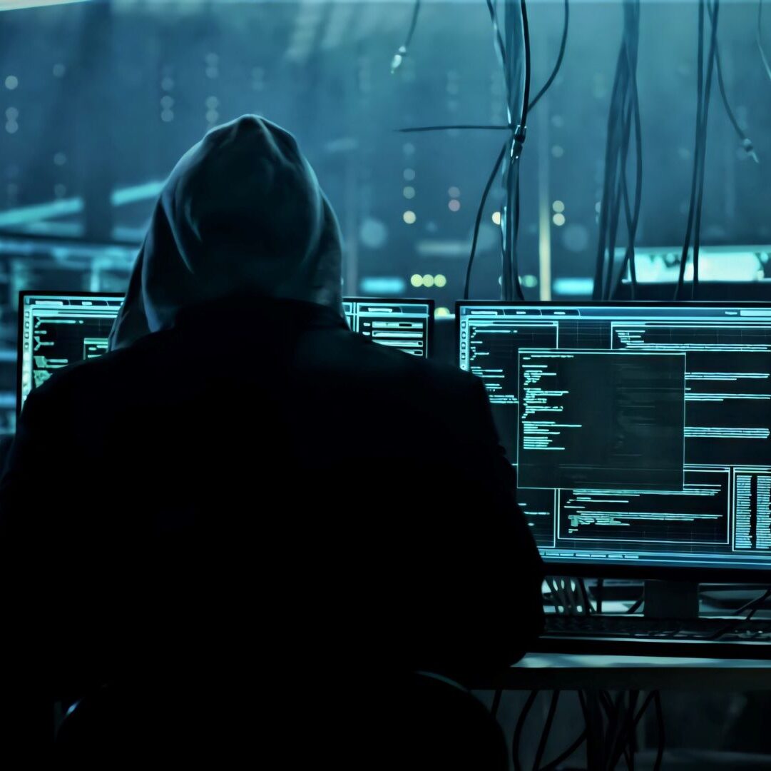 A cybercriminal with his hood up. There are lines of code on two monitors in the background.